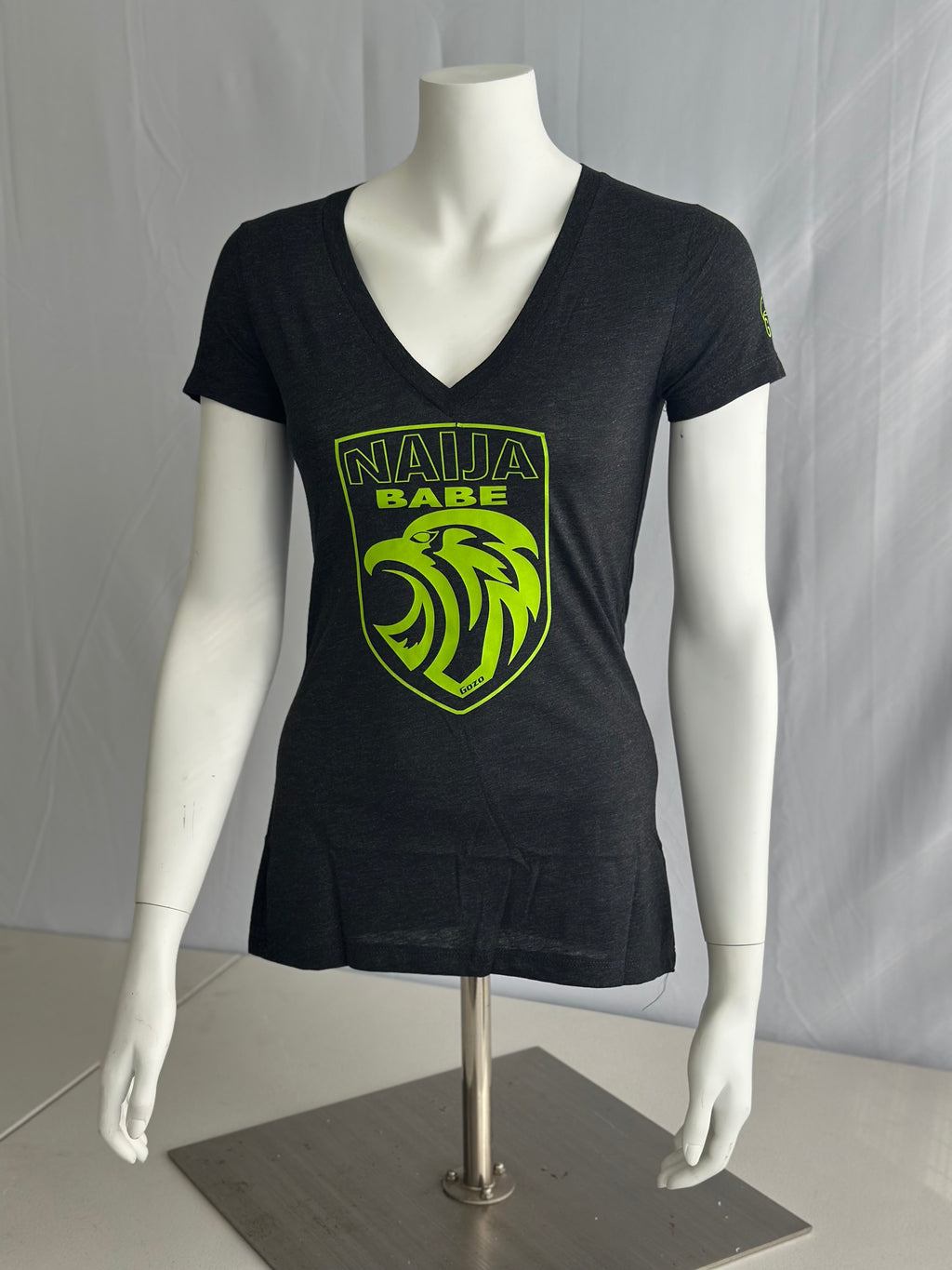 An ash-gray short-sleeve V-neck t-shirt displayed on a white mannequin torso. The shirt features a bright neon green graphic on the front with the text 'NAILA BABE' above a stylized eagle head inside a shield.