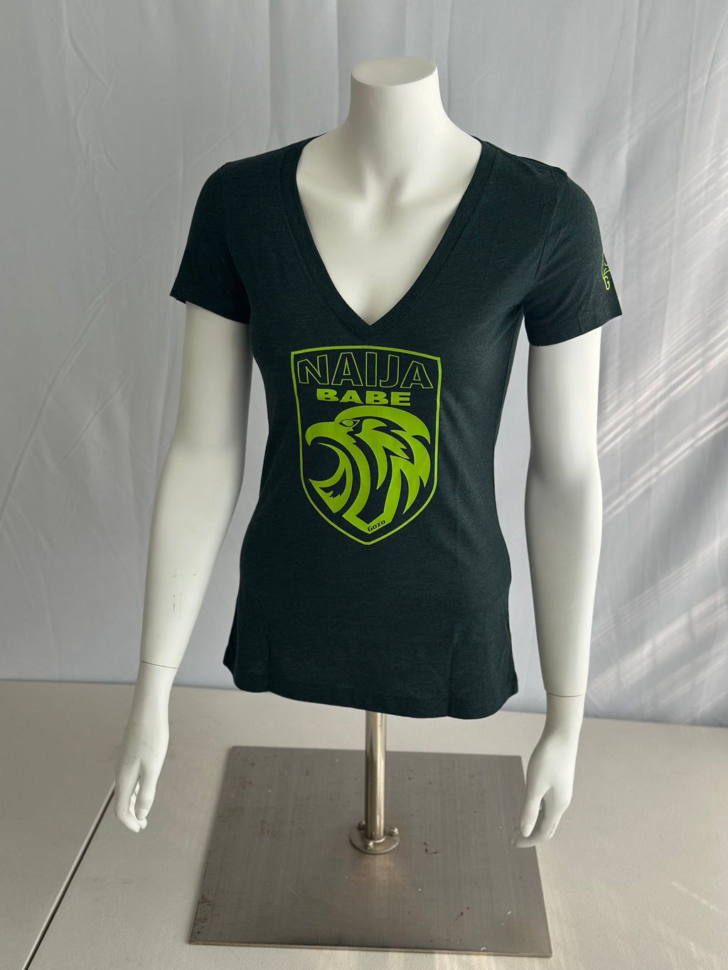 A dark green short-sleeve V-neck t-shirt displayed on a white mannequin torso with arms. The shirt features a bright lime green shield graphic on the chest with a stylized lion head and the text 'NAIJA BABE' in lime green.