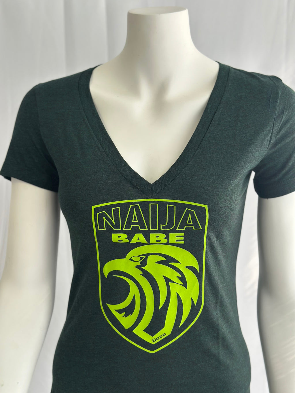 A dark green v-neck t-shirt displayed on a white mannequin torso. The shirt features a bright lime green graphic on the chest with the text 'NAIJA BABE' and a stylized eagle head inside a shield outline.