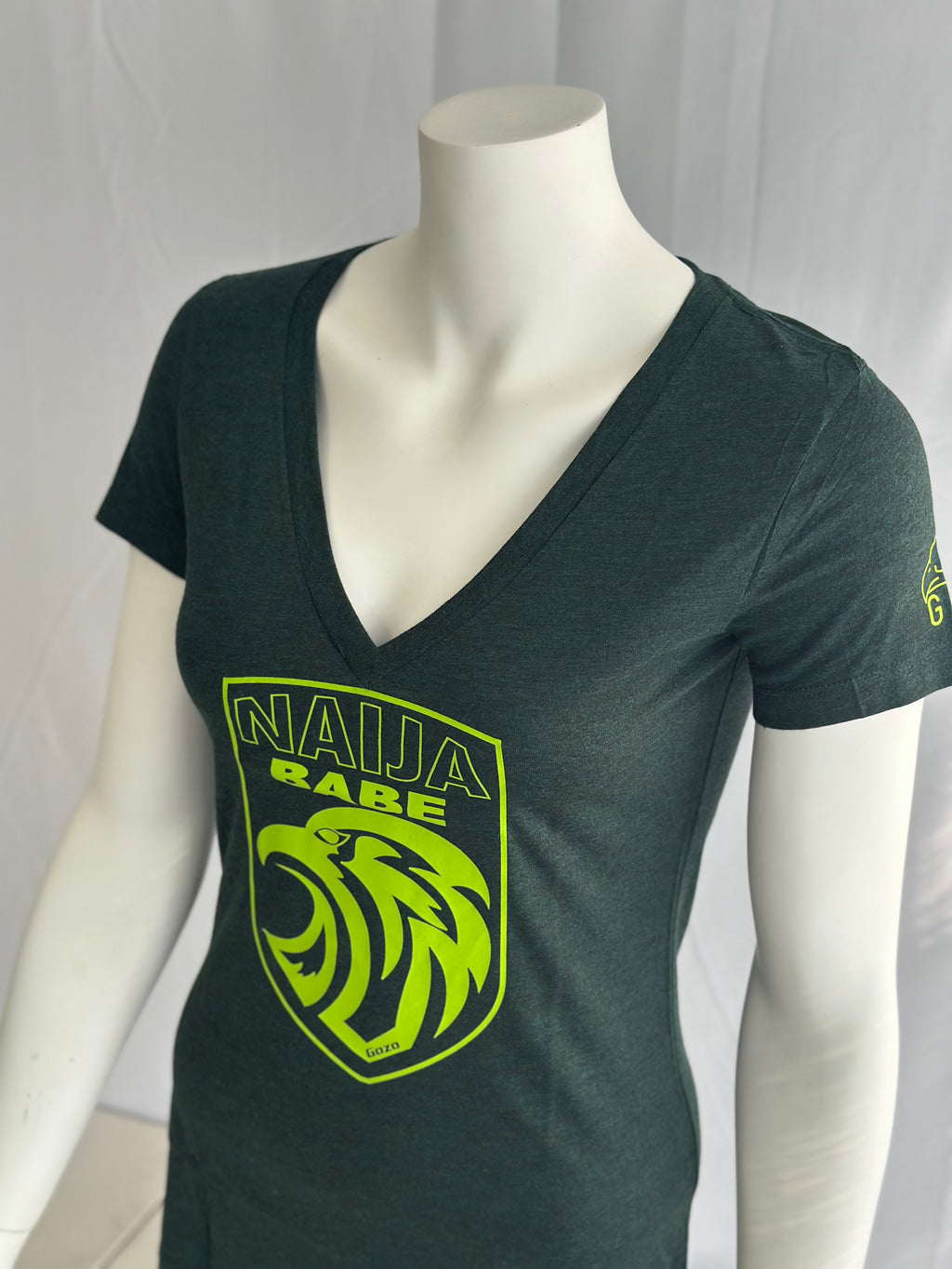 A dark green v-neck t-shirt displayed on a white mannequin torso. The shirt features a bright neon yellow-green graphic on the front with the text 'NAIJA BABE' and a stylized eagle head inside a shield, with a similar neon graphic on the left sleeve.